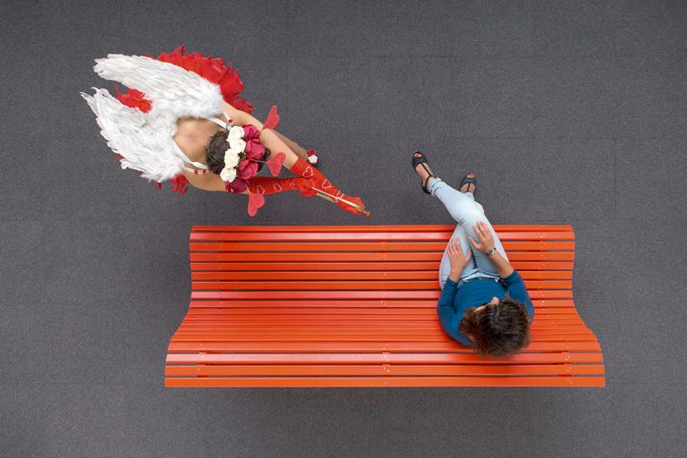 Cupidon avec son arc et ses flèches vise une jeune fille sur un banc rouge.