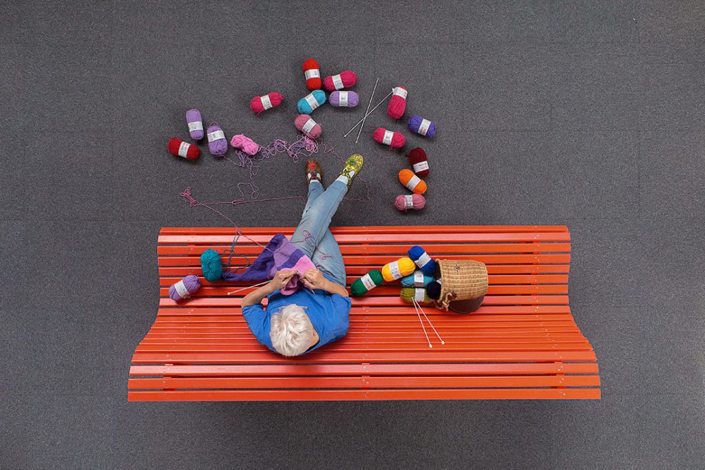 Une grand-mère est assise sur un banc rouge en tricotant des pelotes de laines multicolores.